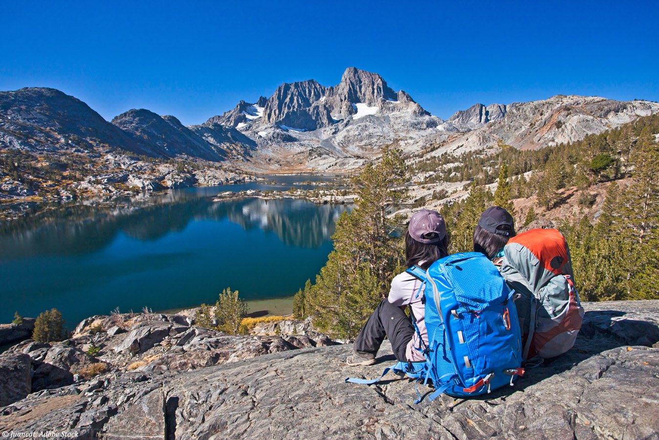 Two girls with backpacks sitting on top of hill, looking at a la